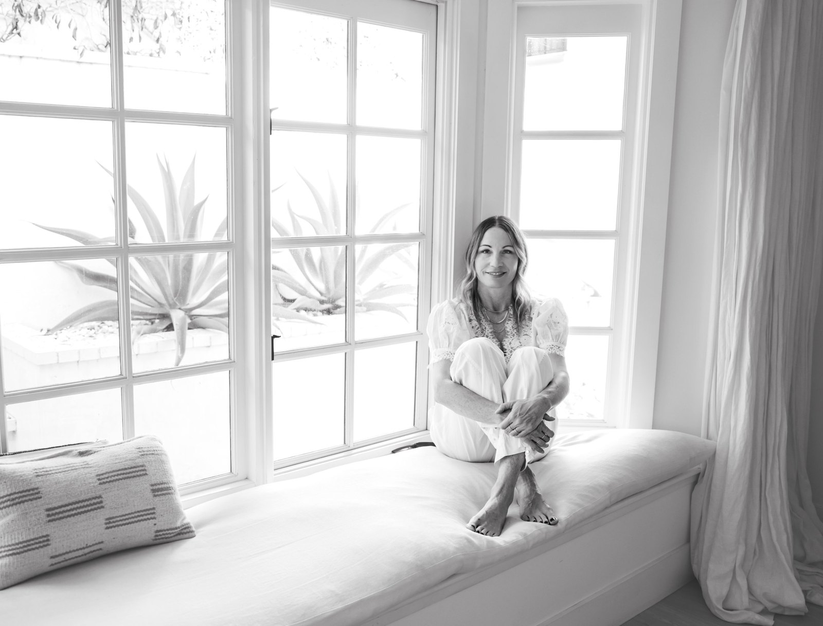 Black and white portrait of Marlene Hennings sitting on a window seat in a white room, agave plants outside