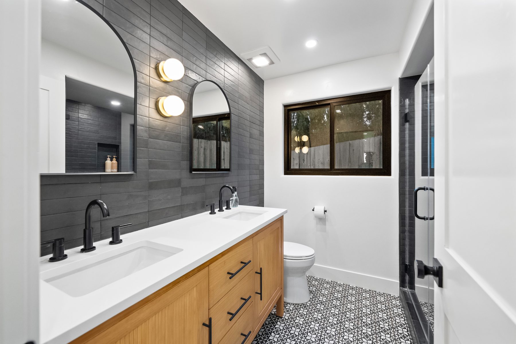 Vista Bahia bath with charcoal tile, arched mirrors, globe sconces, an oak double vanity and a patterned floor