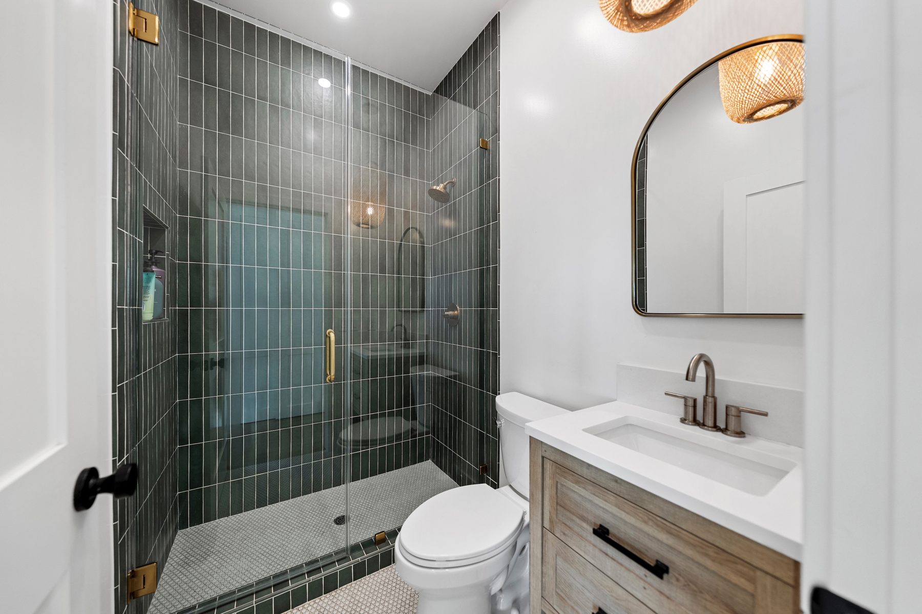 Vista Bahia bath with a green tiled shower, brass hardware, an arched mirror and a woven pendant