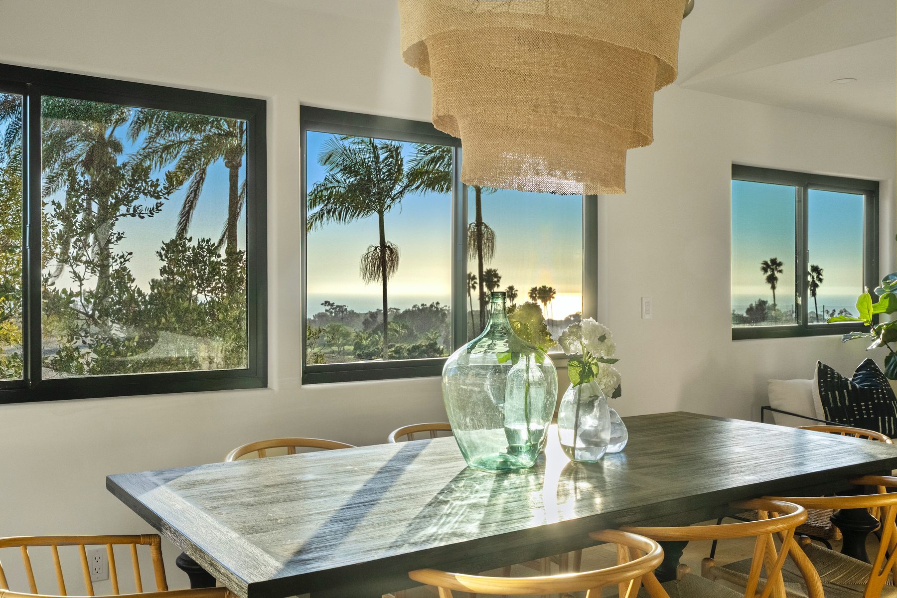Vista Bahia dining room with a dark wood table, wishbone chairs, a tiered woven pendant and ocean views at sunset