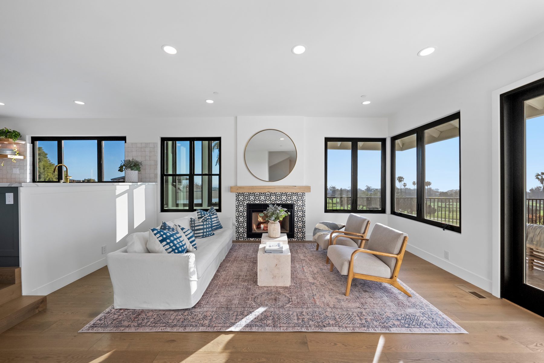 Vista Bahia living room with a white slipcovered sofa, a patterned-tile fireplace under a round mirror, and black-framed windows onto the golf course